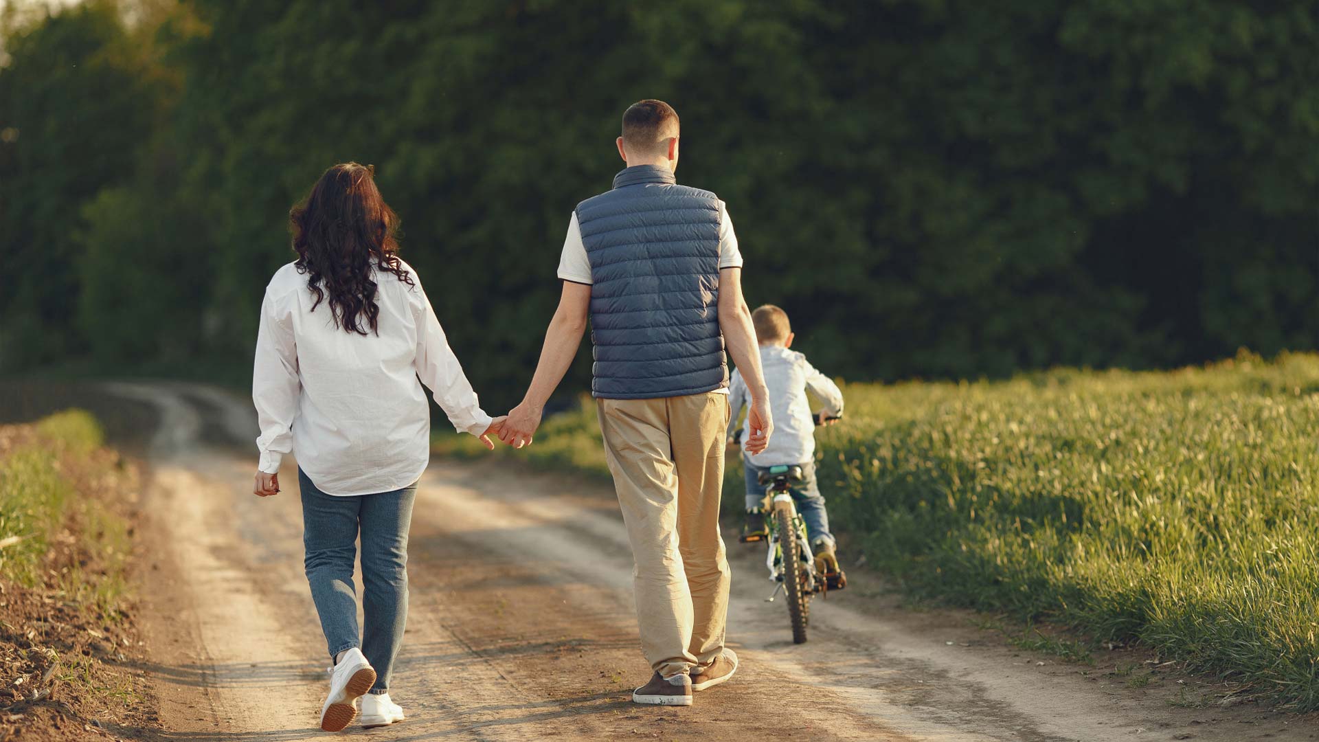 A wife and husband business team with their young child are pictured walking down a dirt road while the child rides a bicycle.