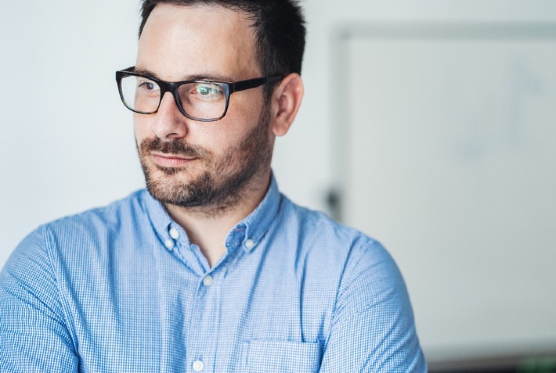 A white man with neutral brown glasses wears a blue button down shirt and stares out the window with a neutral face.