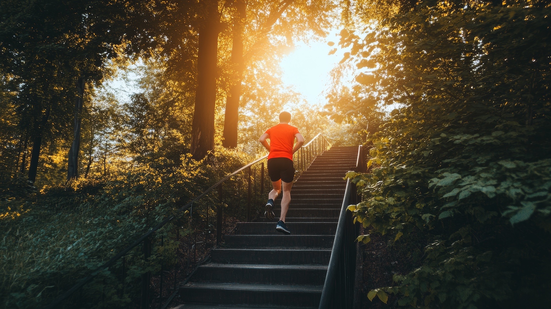 A man runs up the steps outdoors wearing running shorts and a bright read shirt while the sun rises in the background.